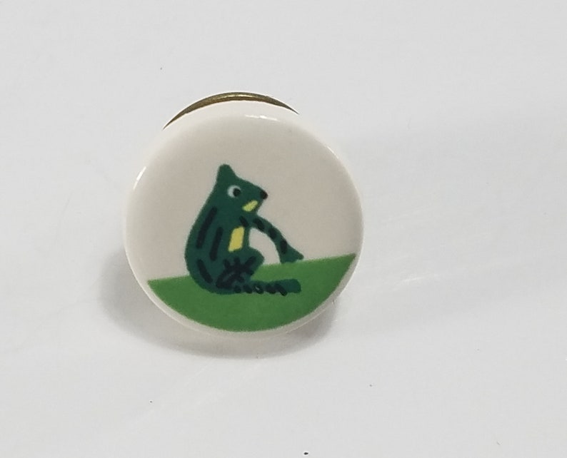 May include: A round ceramic knob with a green frog illustration on a green background. The knob has a gold rim.