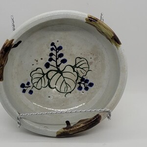 May include: A round, off-white ceramic bowl with a hand-painted floral design. The bowl features a central motif of green leaves and blue berries. Brown and tan accents are painted around the rim. The bowl is suspended by a metal wire.