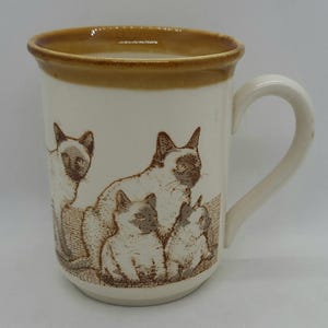 May include: A cream-colored ceramic mug featuring a brown rim and a brown graphic illustration of three Siamese cats. The design depicts one standing cat and two smaller seated cats, all rendered in a detailed, stippled brown ink style.