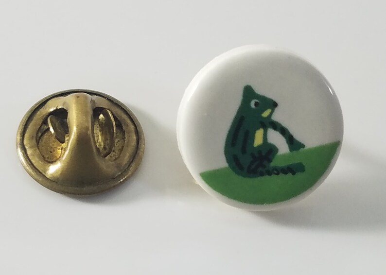 May include: A green frog pin with a white background and a green grass base. The pin has a brass back with a pin closure.