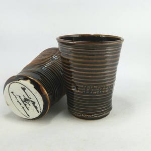 May include: Two handmade ceramic tumblers. The tumblers are dark brown with horizontal ridged detailing and a glossy finish. One tumbler is upright, the other is lying on its side, revealing a white base with a black design.