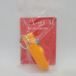 May include: An orange keychain with a silver ring, packaged in a clear plastic bag. The keychain has a small, colorful design. The packaging includes the text "AVON The Gift Collection" on a red background.