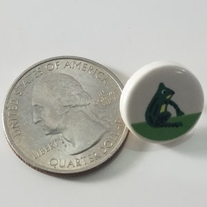 May include: A white ceramic button with a green and black frog illustration. The button is about the size of a US quarter.
