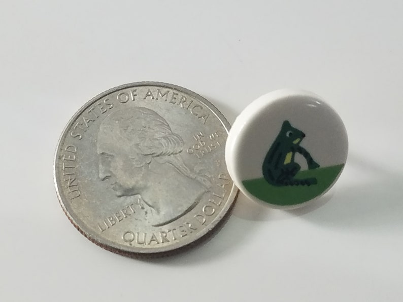 May include: A white ceramic button with a green and black frog illustration. The button is about the size of a US quarter.