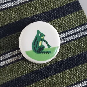 May include: A white ceramic button with a green frog illustration. The frog is sitting on a green leaf and has a yellow belly.