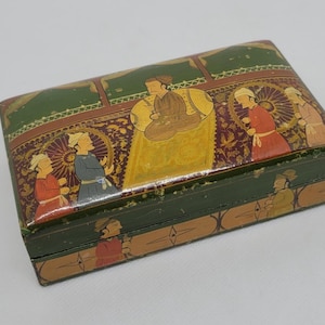 May include: A rectangular, antique wooden box with a hinged lid. The box is painted with a detailed scene featuring figures in traditional attire, set against a backdrop of intricate patterns and gold accents. The box is primarily green with a gold and red design.