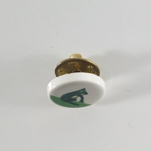 May include: A white ceramic button with a green frog design. The button has a brass shank.