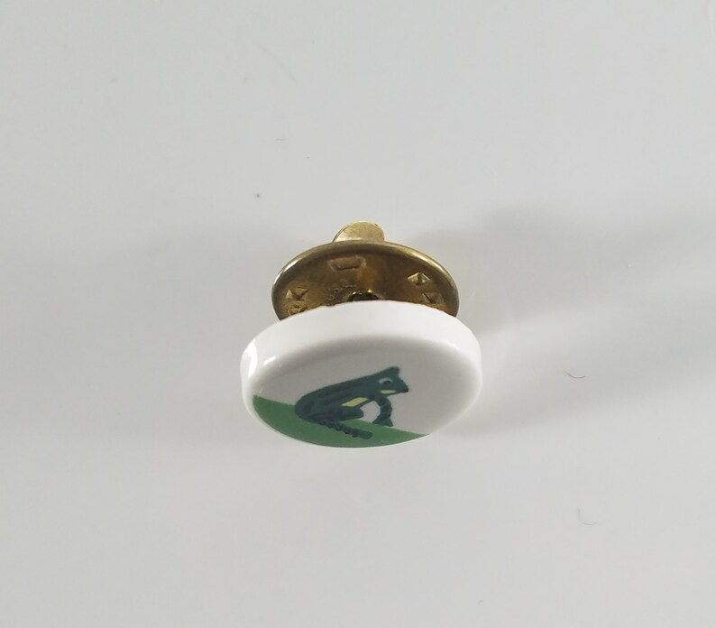 May include: A white ceramic button with a green frog design. The button has a brass shank.