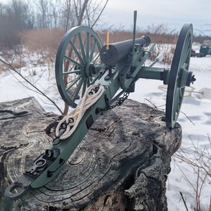 Può includere: Un modello dettagliato di un cannone verde e nero, con grandi ruote a raggi e dettagli in corda. Il cannone è montato su un ceppo di legno stagionato, su uno sfondo innevato.