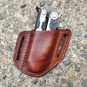 Pode incluir: Um estojo de couro marrom para ferramentas multifuncionais com uma ferramenta multifuncional prateada e preta dentro.