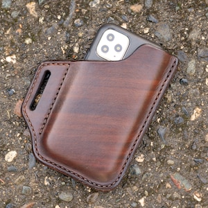 May include: A brown leather phone case with a dark finish. The case has a holster-style design with a belt loop and visible stitching. A smartphone is partially visible inside the case. The case is designed to protect the phone.