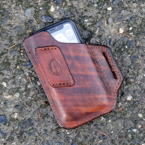 May include: A brown leather phone holster with a stitched design. The holster is designed to hold a smartphone with a screen size of approximately 6 inches.