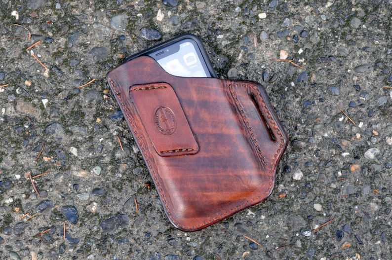 May include: A brown leather phone holster with a stitched design. The holster is designed to hold a smartphone with a screen size of approximately 6 inches.