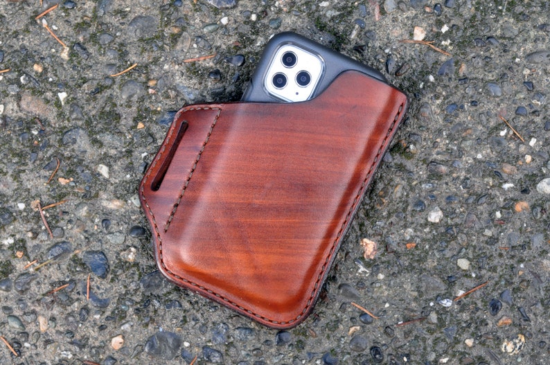 May include: A brown leather phone case with a stitched edge. The case has a belt loop and a slot for a phone. A silver phone with a black case is inside the leather case.