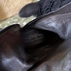 May include: Black leather boots with fringe detailing. The boots have a pointed toe and a zipper closure. The interior of the boot features the brand name "DONALD J PLINER" and "MADE IN ITALY".