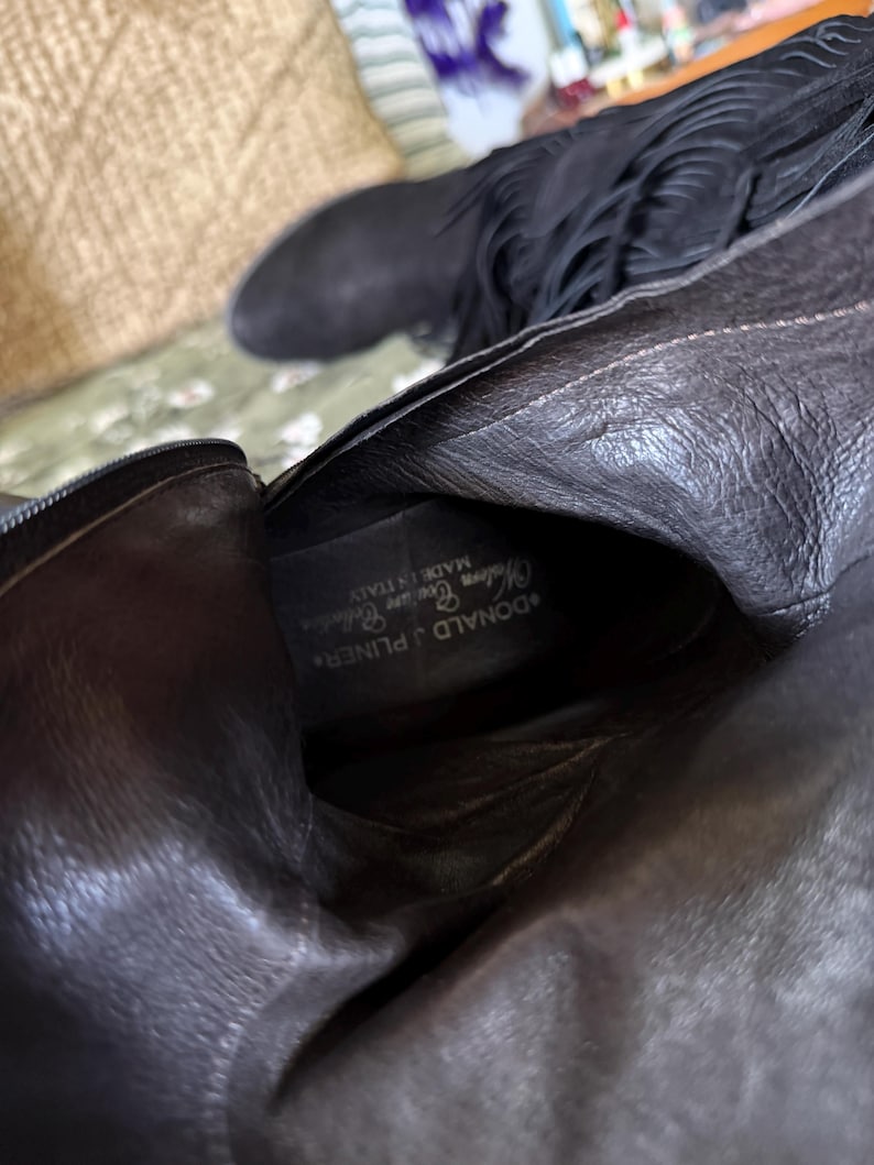May include: Black leather boots with fringe detailing. The boots have a pointed toe and a zipper closure. The interior of the boot features the brand name "DONALD J PLINER" and "MADE IN ITALY".