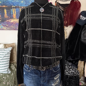 May include: A black sweater with a gray plaid pattern is displayed on a mannequin. The sweater is paired with a denim skirt and a studded belt. A silver necklace with a circular pendant is visible.