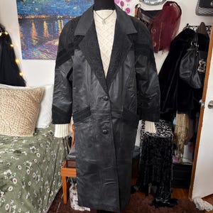 May include: A black leather coat with a textured collar and decorative panels on the shoulders. The coat has a button-down front and is displayed on a mannequin. The coat is paired with a cream-colored sweater.