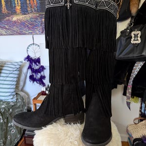 May include: A pair of black suede knee-high boots with fringe detailing. The boots have a decorative beaded band at the top and a small heel. The boots are displayed on a fluffy white surface.