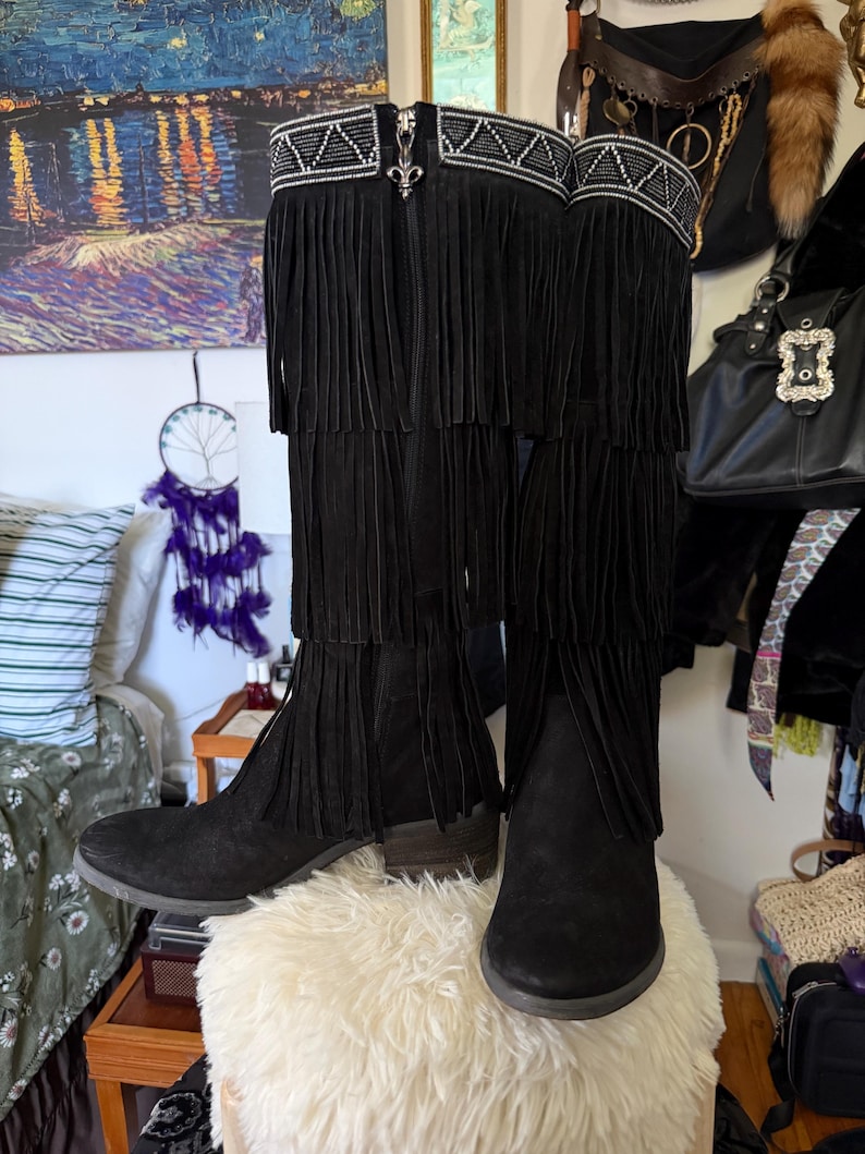 May include: A pair of black suede knee-high boots with fringe detailing. The boots have a decorative beaded band at the top and a small heel. The boots are displayed on a fluffy white surface.