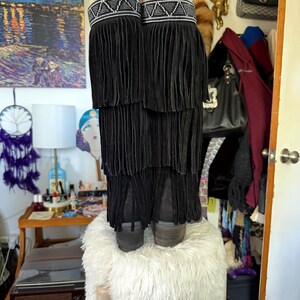 May include: Black suede boots with layered fringe detailing. The boots have a decorative band at the top with white and silver beading. The boots are displayed on a white, fluffy stool. The boots have a stacked heel.