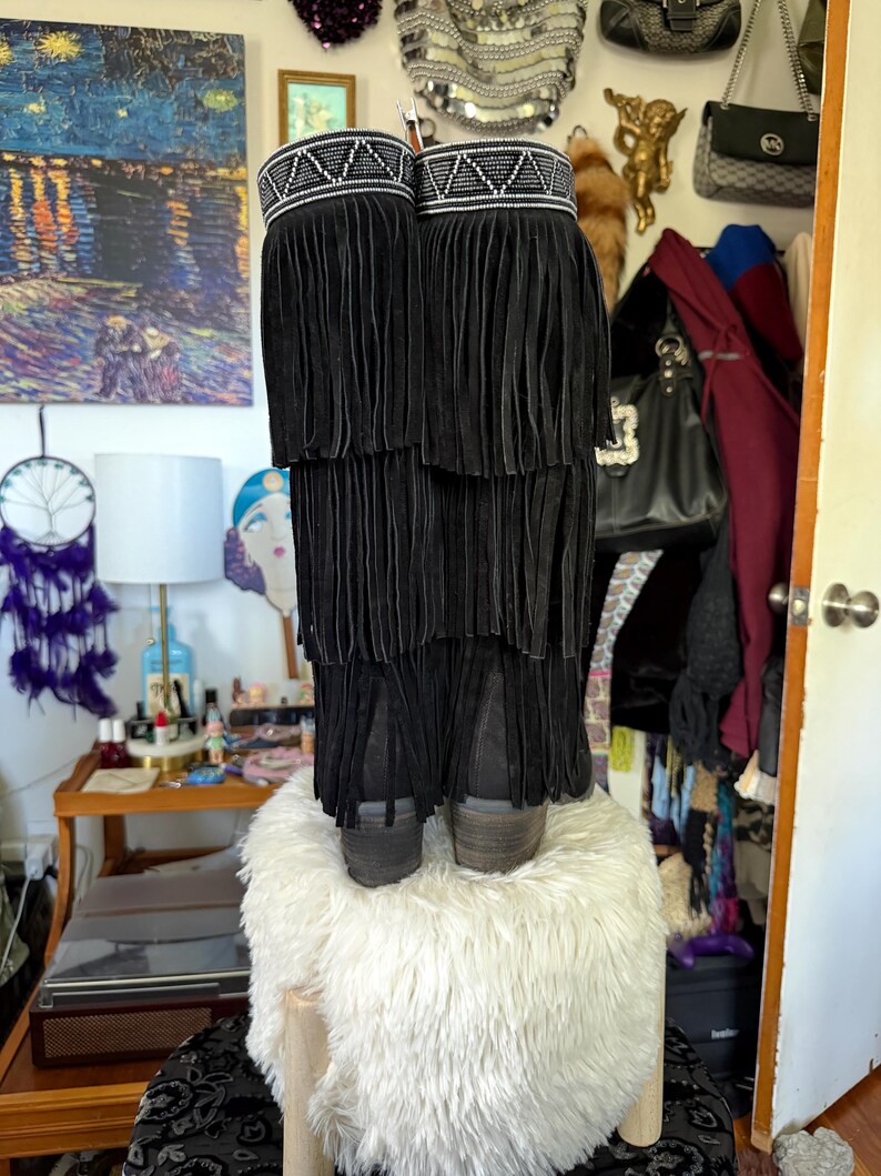 May include: Black suede boots with layered fringe detailing. The boots have a decorative band at the top with white and silver beading. The boots are displayed on a white, fluffy stool. The boots have a stacked heel.