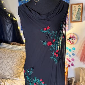 May include: Black sleeveless dress with a cowl neck, adorned with embroidered red roses and green leaves. The floral design is accented with black beads. A gold necklace with a small pendant is visible on the mannequin.