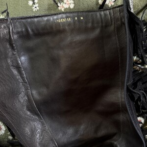 May include: Black leather knee-high boot with fringe detail. The interior shows the text "IDENISE 8 M" in gold lettering. The boot has a zipper and is set against a floral patterned background.