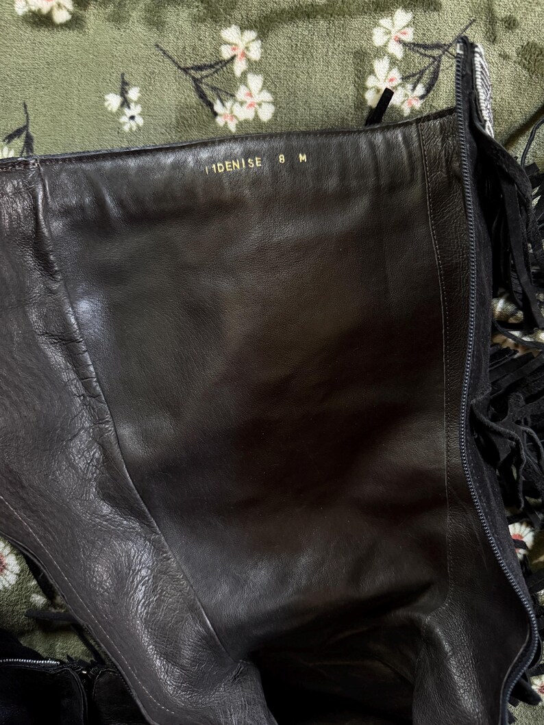 May include: Black leather knee-high boot with fringe detail. The interior shows the text "IDENISE 8 M" in gold lettering. The boot has a zipper and is set against a floral patterned background.