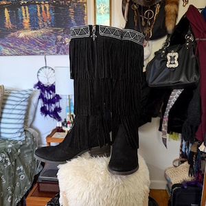 May include: Black suede boots with fringe detailing. The boots have a decorative silver trim at the top and a zipper closure. The boots are displayed on a white, fluffy ottoman. The boots are a stylish fashion item.