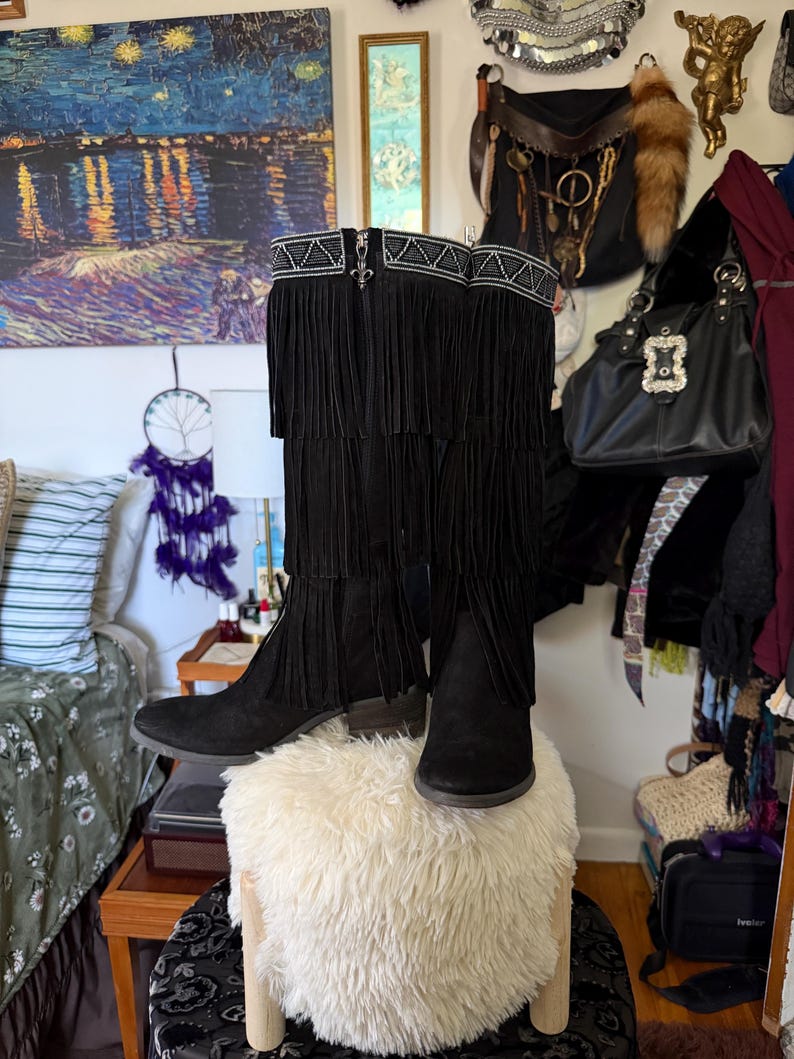 May include: Black suede boots with fringe detailing. The boots have a decorative silver trim at the top and a zipper closure. The boots are displayed on a white, fluffy ottoman. The boots are a stylish fashion item.
