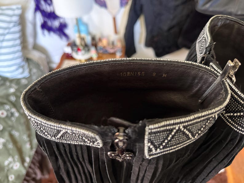 May include: Close-up of a black leather boot with fringe and a decorative beaded trim around the top. The interior shows the text "DENISE 8 M". A silver zipper with a decorative pull is visible.