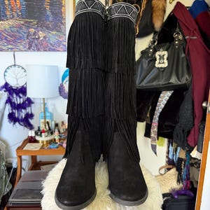 May include: Black suede boots with multiple layers of fringe and a decorative beaded band at the top. The boots are knee-high and have a low heel. The boots are displayed on a fluffy white surface.