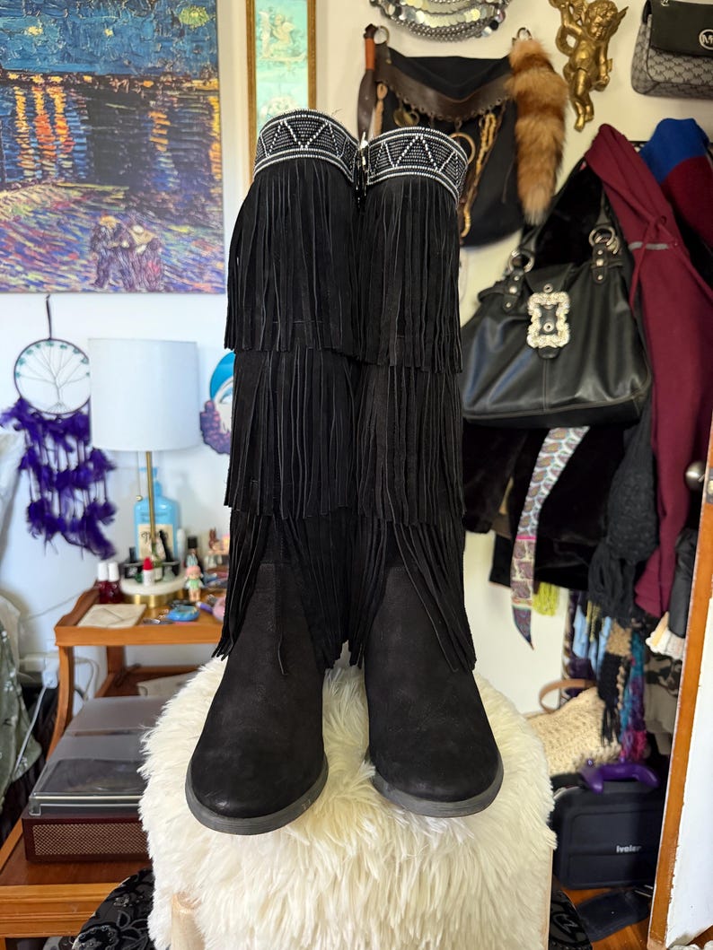 May include: Black suede boots with multiple layers of fringe and a decorative beaded band at the top. The boots are knee-high and have a low heel. The boots are displayed on a fluffy white surface.