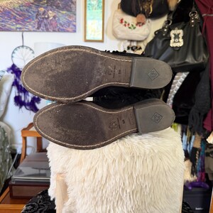 May include: A pair of black leather boots with worn brown soles. The soles are marked with a logo and size information. The boots are placed on a fluffy, white, round stool.