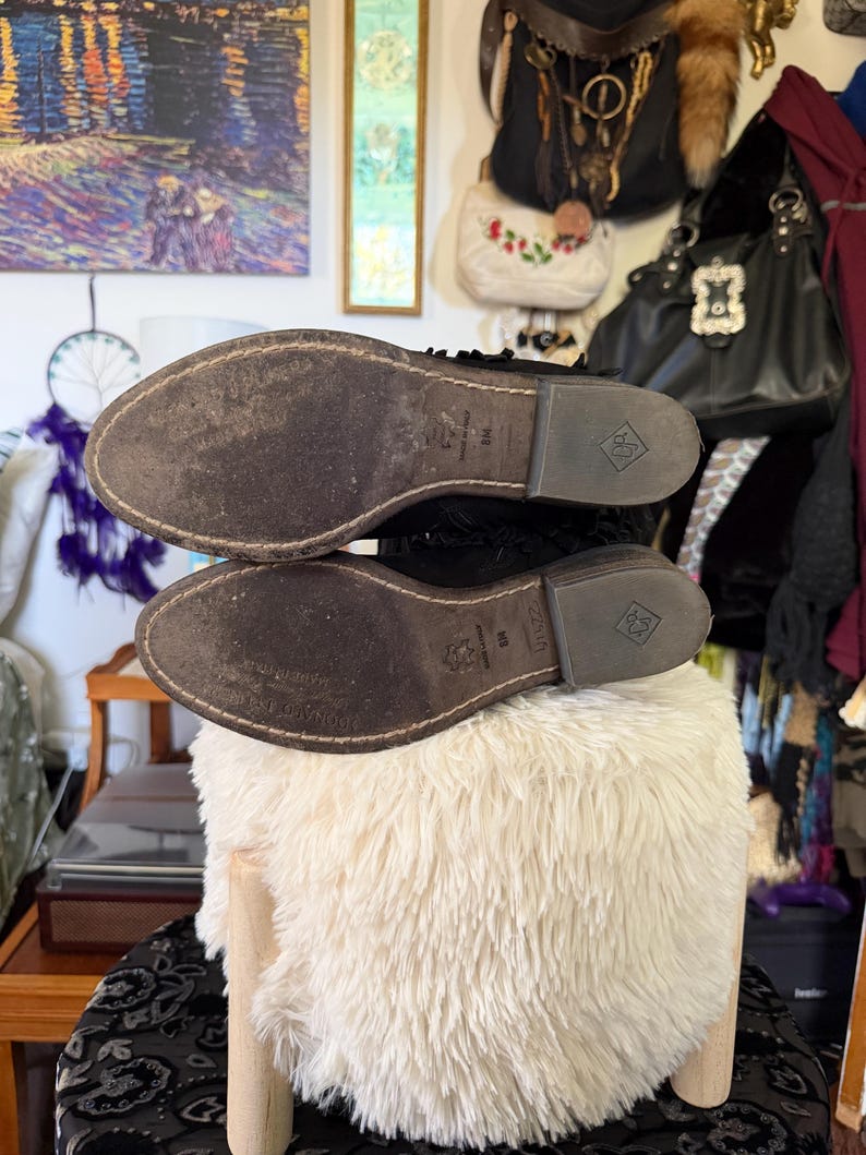 May include: A pair of black leather boots with worn brown soles. The soles are marked with a logo and size information. The boots are placed on a fluffy, white, round stool.
