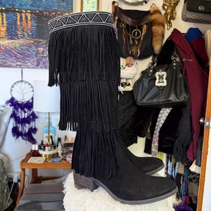 May include: Black suede knee-high boots with fringe detailing. The boots feature a decorative beaded band at the top and a stacked heel. The fringe cascades down the front of the boot in multiple layers, creating a bohemian style.