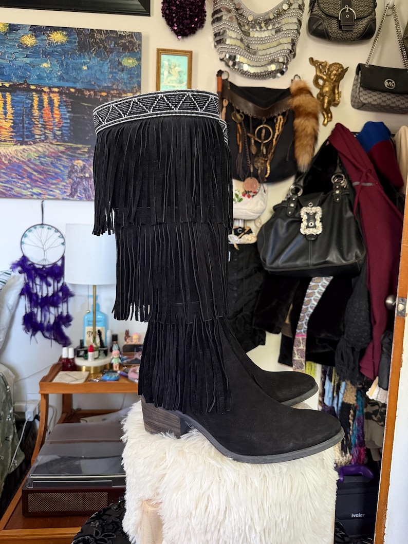 May include: Black suede knee-high boots with fringe detailing. The boots feature a decorative beaded band at the top and a stacked heel. The fringe cascades down the front of the boot in multiple layers, creating a bohemian style.