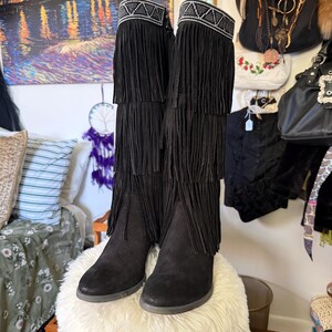May include: A pair of black suede boots with fringe detailing. The boots have a decorative beaded band at the top and multiple layers of fringe cascading down the shaft. The boots are displayed on a fluffy, white ottoman.