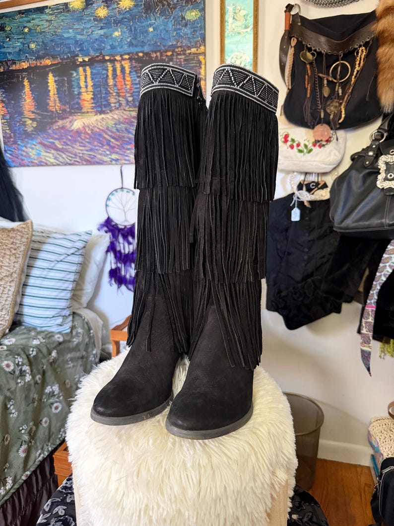 May include: A pair of black suede boots with fringe detailing. The boots have a decorative beaded band at the top and multiple layers of fringe cascading down the shaft. The boots are displayed on a fluffy, white ottoman.