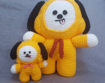 May include: Two crocheted plush toys shaped like dogs. The larger dog is yellow with a white face and black ears. The smaller dog is yellow with a white face and black ears. Both dogs have a red ribbon around their necks.