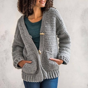 May include: A grey crocheted cardigan with pockets and a button closure. The cardigan is made with a textured stitch and has a relaxed fit.