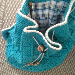 Puede incluir: Una bolsa de crochet azul turquesa con ribete de crochet blanco y cierres de bot&oacute;n de madera. La bolsa tiene un forro de tela a cuadros.
