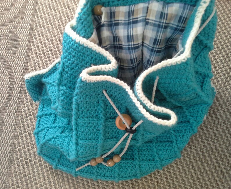 Puede incluir: Una bolsa de crochet azul turquesa con ribete de crochet blanco y cierres de bot&oacute;n de madera. La bolsa tiene un forro de tela a cuadros.