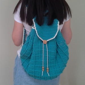 May include: A turquoise crocheted backpack with white trim and wooden button closures. The backpack has a drawstring closure and is worn by a person.