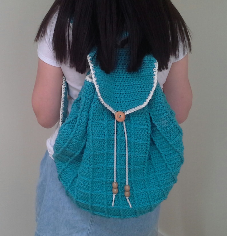 Puede incluir: Una mochila de crochet turquesa con ribetes blancos y cierres de bot&oacute;n de madera. La mochila tiene un cierre de cord&oacute;n y la lleva puesta una persona.