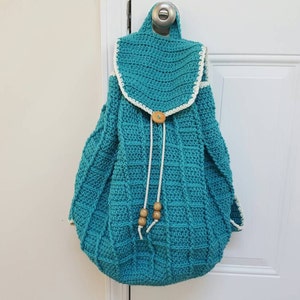 Puede incluir: Una mochila de crochet azul turquesa con ribete de crochet blanco y cierre de bot&oacute;n de madera. La mochila tiene cierre de cord&oacute;n y cuentas de madera en los cordones.