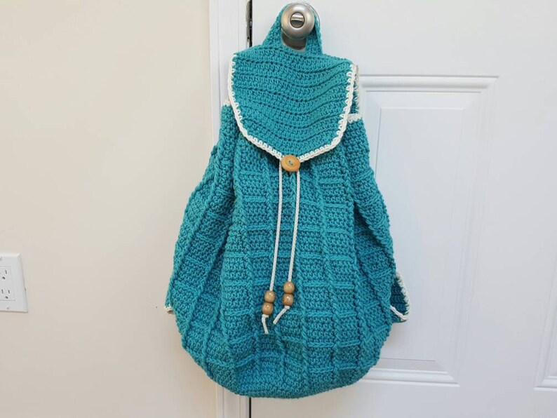 Puede incluir: Una mochila de crochet azul turquesa con ribete de crochet blanco y cierre de bot&oacute;n de madera. La mochila tiene cierre de cord&oacute;n y cuentas de madera en los cordones.