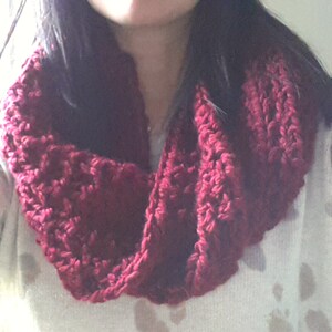 May include: A burgundy-colored, chunky knit infinity scarf. The scarf is made of a thick yarn, creating a textured appearance. The knitted loops are visible, adding to the handmade aesthetic. The scarf is designed to be worn around the neck, offering warmth and style.