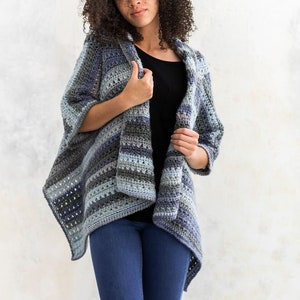 Puede incluir: Un cárdigan de crochet a rayas azules y grises con un frente largo y abierto. El cárdigan se lleva sobre una camisa negra y unos vaqueros azules.